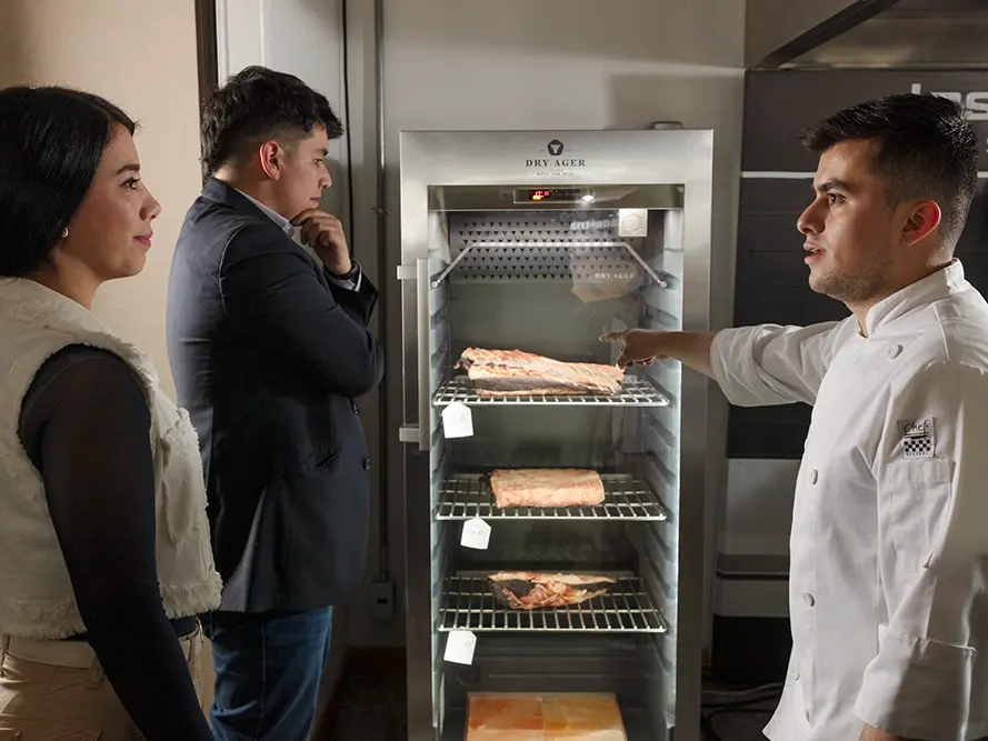 Un chef está mostrando un aparato de maduración en seco con carne a dos personas.